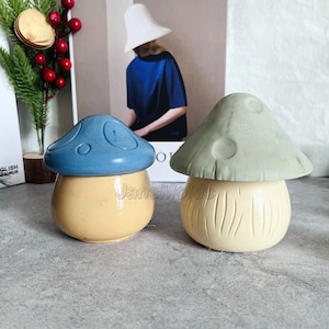 Puede incluir: Dos recipientes de cerámica con forma de hongo con tapas. Un recipiente es azul claro con una base beige, y el otro es verde claro con una base beige.