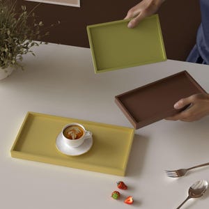 Op de afbeelding: Rechthoekige trays in olijfgroen, bruin en geel. Eén gele tray bevat een witte kop en schotel met een latte. Aardbeien en bestek liggen ook op tafel. De trays worden door een persoon vastgehouden.