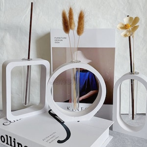 Może przedstawiać: Trzy białe ceramiczne wazony z szklanymi rurkami do przechowywania suszonych kwiatów. Wazony mają różne kształty: kwadratowy, okrągły i owalny. Wazony stoją na książce z napisem "Collins" na okładce.