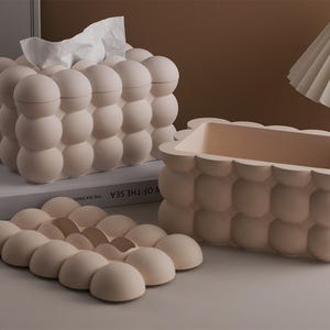 Puede incluir: Un conjunto de tres elementos decorativos beige con diseño de burbujas. El conjunto incluye un dispensador de pañuelos, un recipiente rectangular y una bandeja. El dispensador de pañuelos contiene pañuelos blancos. Los elementos están dispuestos sobre una superficie blanca.