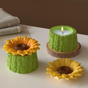 Op de afbeelding: Drie decoratieve items met zonnebloemthema. Eén is een kandelaar met een brandende kaars, geplaatst op een houten onderzetter. Een andere is een zonnebloemvormige container. De derde is een zonnebloemvormig schaaltje.