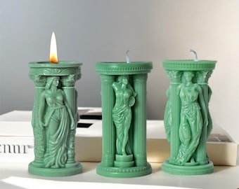 Puede incluir: Tres velas de pilar verdes con una figura femenina esculpida en cada una. Las velas están encendidas y tienen una llama en la parte superior.