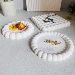 Peut inclure: Trois moules ronds en silicone blanc avec des bosses surélevées autour des bords. Les moules sont destinés à la fabrication de plats à bijoux. Un moule contient des fleurs séchées, un autre une paire de boucles d'oreilles dorées et le troisième un collier noir.