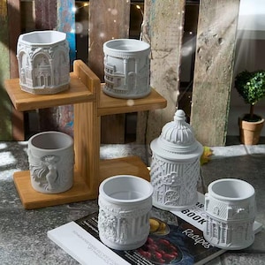 May include: A collection of five white, cylindrical, and hexagonal containers featuring detailed architectural and sculptural reliefs. One container has a domed lid, while the others are open-topped. They are displayed on a wooden stand and a book.