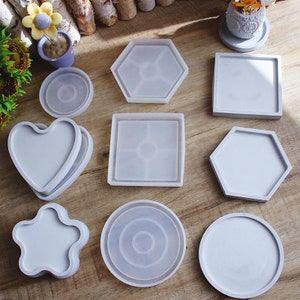 May include: A selection of grey silicone moulds in diverse shapes, such as a heart, star, hexagon, square, and circle. These moulds are intended for crafting and creating decorative items. The shapes are arranged on a wooden surface.