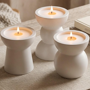 May include: Three white candle holders with smooth, matte finishes and distinct geometric shapes, each containing a lit candle with a visible flame. The holders vary in height and silhouette, featuring rounded, tapered, and stacked circular forms.
