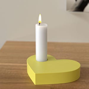 Op de afbeelding: Een witte kaars met een gele vlam in een gele hartvormige kandelaar. De kandelaar staat op een houten oppervlak. De kaars is aangestoken.