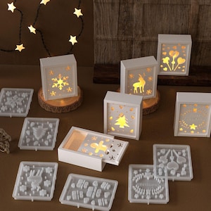 May include: Several white, illuminated Christmas-themed boxes with cut-out designs like snowflakes, a reindeer, a Christmas tree, and hearts. The boxes are on wooden rounds, with crafting molds nearby. A string of star-shaped lights hangs in the background.