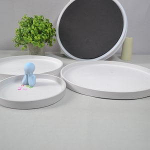 Könnte beinhalten: Drei weiße Keramiktabletts in verschiedenen Größen, mit einer kleinen blauen Figur, die auf dem kleinsten Tablett sitzt. Die Figur hält ihre Hände vor ihrem Gesicht zusammen. Um die Figur herum liegen kleine bunte Herzen.