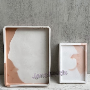 Op de afbeelding: Twee rechthoekige trays gemaakt van beton met een wit en roze marmerpatroon. De grotere tray is links en de kleinere rechts.