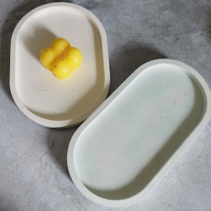 May include: Two oval-shaped concrete trays. One tray is white and the other is light blue with a white rim. The white tray has a yellow wax candle in the shape of a flower.