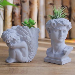 Könnte beinhalten: Zwei weiße Keramik-Pflanzgefäße in Form einer Engels-Skulptur und einer Männerbüste. Der Engel-Pflanztopf hat eine kleine Sukkulente drin. Der Büsten-Pflanztopf hat eine kleine Luftpflanze drin.