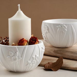 May include: Two white ceramic bowls with embossed floral designs. One bowl holds a white candle, dried flowers, and decorative elements. The other bowl is empty. The bowls are on a wooden surface, creating a decorative display.