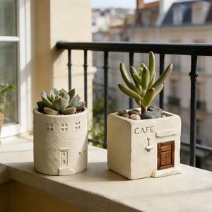 May include: Two beige succulent planters. One is cylindrical with a door and window cutouts. The other is square with a door and the word "CAFE". Both are on a ledge with a balcony in the background. The plants are green.