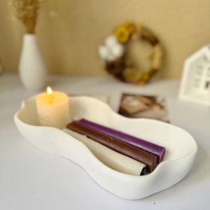 Könnte beinhalten: Weiße Keramikschale mit wellenförmigem Rand, die eine weiße Kerze und drei Stumpenkerzen enthält. Die Kerzen sind lila, braun und weiß.