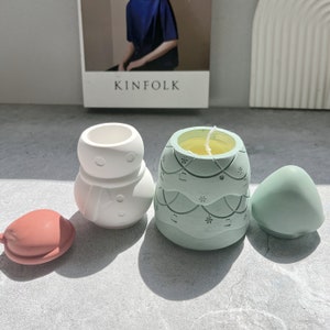 Könnte beinhalten: Ein Set aus vier dekorativen Kerzenhaltern in Form eines Schneemanns, eines Weihnachtsbaums und zwei Ornamenten. Der Weihnachtsbaum-Kerzenhalter ist grün mit einem weißen Schneeflockenmuster und hat eine gelbe Kerze im Inneren. Der Schneemann-Kerzenhalter ist weiß mit einem schwarzen Hut und Knöpfen. Die Ornamente sind rot und grün.