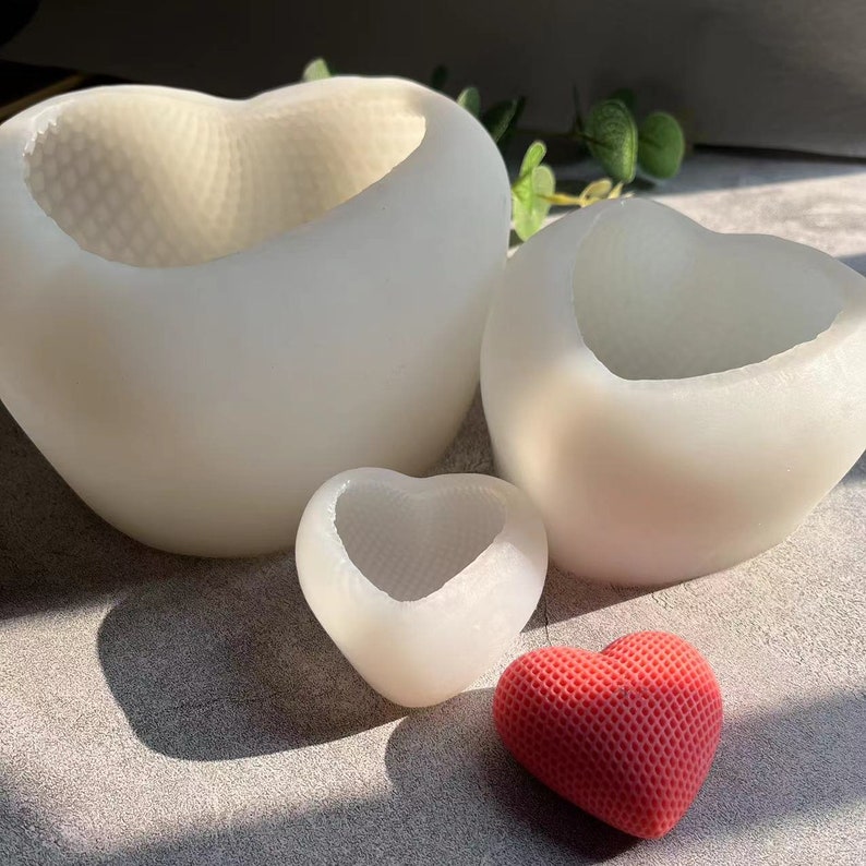 以下が含まれることがあります： 3つの異なるサイズの白いハート型のシリコンモールド。最も小さいモールドがフォーカスされ、赤いハート型のオブジェクトが前景にあります。