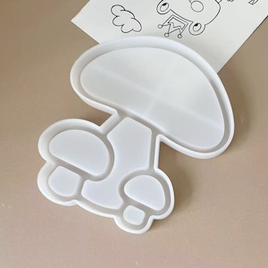 May include: A white mushroom-shaped mold for crafting. The mold has a large, rounded top and a stem with several smaller, rounded sections. The design is smooth and clean, perfect for resin or other craft projects. A drawing of a cartoon frog is in the background.