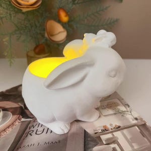 Könnte beinhalten: Ein weißer Keramik-Kerzenhalter in Hasenform mit warmem gelbem Schein von innen. Der Hase hat lange Ohren und sitzt auf einem Buch. Dekorative Elemente sind im Hintergrund sichtbar.