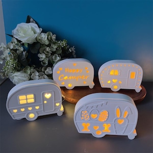 Puede incluir: Cuatro luces nocturnas blancas con forma de caravana, con diseños recortados e iluminación amarilla cálida. Una luz tiene el texto "Happy Camper". Otros diseños incluyen corazones, ventanas y un conejito. Las luces están sobre una superficie de madera.
