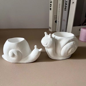 May include: Two white ceramic snail planter pots. The larger snail has a wide, open shell, while the smaller snail has a more closed shell. Both snails have a cute, cartoon-like face.