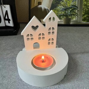 May include: White wooden candle holder shaped like a house with a heart cutout. The holder has a small, round, pink candle inside.