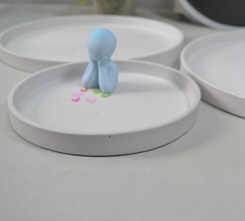 Pu&ograve; includere: Un vassoio in ceramica bianca con una statuetta blu chiaro seduta al centro. La statuetta &egrave; seduta con le mani sul mento e ha un piccolo mucchio di cuori colorati davanti a s&eacute;.