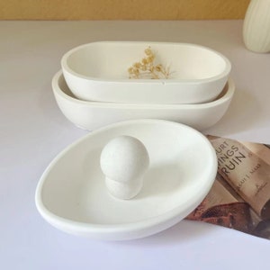 Op de afbeelding: Witte ovale keramische schaal met een kleinere witte ovale keramische schaal erin. De kleinere schaal heeft een witte keramische knop in het midden. De schalen staan op een wit oppervlak.