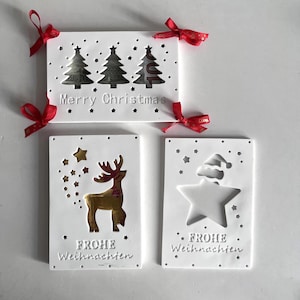 以下が含まれることがあります： 赤いリボンをあしらった白いクリスマスカードが3枚。最初のカードにはクリスマスツリーが3本と「メリークリスマス」の文字が書かれている。2枚目のカードにはトナカイと「Frohe Weihnachten」の文字が書かれている。3枚目のカードには星と「Frohe Weihnachten」の文字が書かれている。