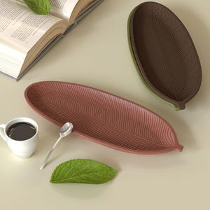 Pode incluir: Duas travessas em forma de folha em tons de castanho e verde. Também estão presentes uma chávena de café branca com café preto, uma colher pequena, uma folha verde e um livro aberto. As travessas têm um design texturizado e canelado.