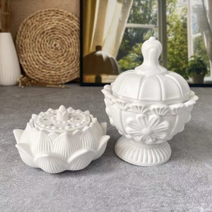 以下が含まれることがあります： 白いセラミック製の香炉が2つ。1つは、小さな蓋と線香用の穴が付いた蓮の花の形をしています。もう1つは、蓋付きの丸い容器で、花柄のデザインが施されています。