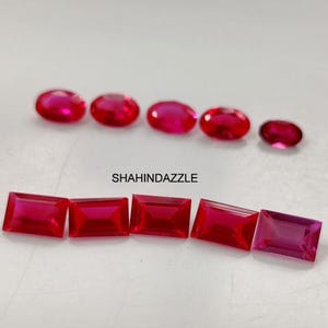 May include: A selection of bright red gemstones, featuring oval and rectangular shapes, are presented on a white background. The gemstones are arranged in two rows, with the text "SHAHINDAZZLE" visible in the middle.