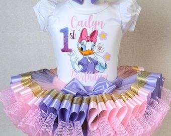 May include: A white bodysuit with Daisy Duck graphic and the text "Cailyn 1st Birthday" paired with a layered tutu skirt in pink, purple, and gold. A matching bow headband completes the outfit.