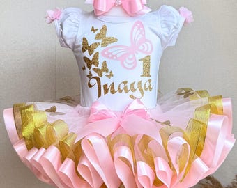 Puede incluir: Un conjunto de tutú rosa y dorado para el primer cumpleaños de un bebé. La parte superior blanca presenta gráficos de mariposas doradas, el número 1 y el nombre "Inaya". El tutú tiene cintas rosas y doradas y un lazo a juego.