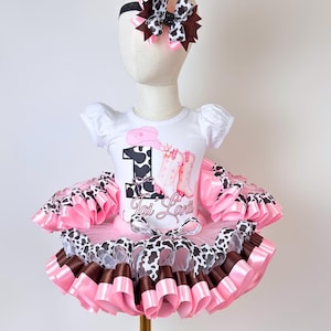 May include: A white short-sleeved bodysuit featuring a graphic of the number one, a pink cowboy hat, and pink boots with cow print accents, paired with a multi-layered pink, brown, and cow print tutu and a matching hair bow on a headband.