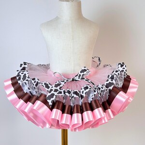 May include: A layered tulle tutu skirt featuring a light pink base with alternating ribbon trim in a black and white cow print pattern, solid dark brown, and solid light pink, tied with a decorative ribbon bow at the center front waist.