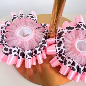 May include: Two circular hair accessories featuring a layered design of pink satin ribbon and black and white cow print ribbon. Each piece is accented with a gathered pink tulle ruffle around the center opening, creating a full, textured appearance.