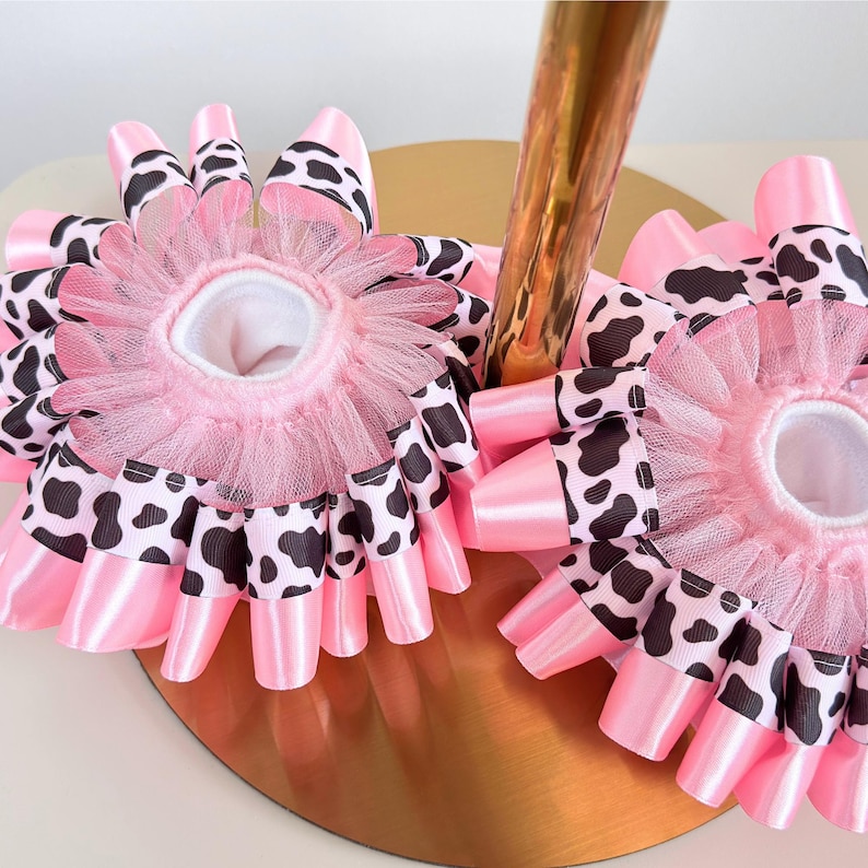 May include: Two circular hair accessories featuring a layered design of pink satin ribbon and black and white cow print ribbon. Each piece is accented with a gathered pink tulle ruffle around the center opening, creating a full, textured appearance.