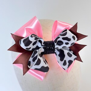 May include: A multi-layered hair bow featuring bright pink satin ribbon, dark brown ribbon points, and a central white ribbon with a black cow print pattern. The center of the bow is secured with a black glittery fabric band for a textured finish.