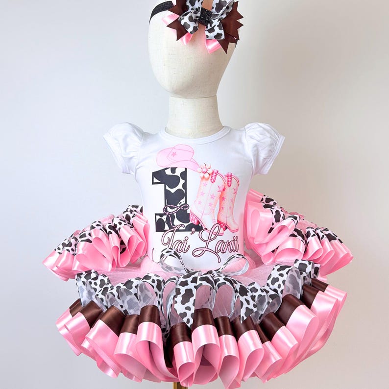 May include: A white short-sleeved bodysuit featuring a graphic of a pink cowboy hat, pink boots, and the number 1 with a cow print pattern. The name Tai Lani is printed below. A layered tutu skirt in pink, brown, and cow print, plus a matching hair bow.