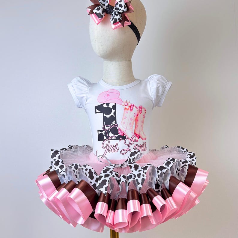 May include: A white short-sleeved bodysuit featuring a black cow print number one, a pink cowboy hat, pink boots, and the name Jai Lani. The outfit includes a layered tutu skirt with pink, brown, and cow print ribbons, and a matching hair bow.