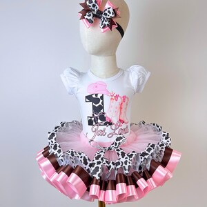 May include: A white short-sleeved bodysuit featuring a cow print number one, a pink cowboy hat, and pink boots graphic with the name Jai Lani. The outfit includes a layered pink, brown, and cow print tutu skirt and a matching hair bow headband.