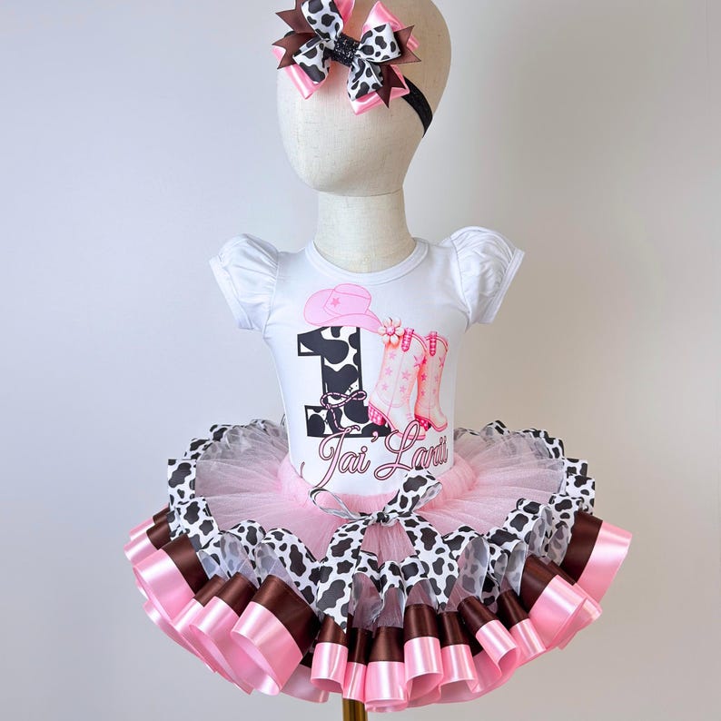 May include: A white short-sleeved bodysuit featuring a cow print number one, a pink cowboy hat, and pink boots graphic with the name Jai Lani. The outfit includes a layered pink, brown, and cow print tutu skirt and a matching hair bow headband.