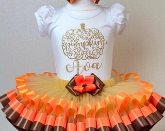 May include: A white short-sleeved bodysuit with a gold glitter pumpkin design and the name "Ava". The outfit includes a tutu skirt with orange, brown, and yellow ribbons, and a matching bow headband. A festive fall-themed outfit.