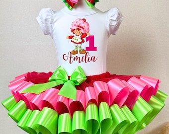May include: A white short-sleeved bodysuit with a Strawberry Shortcake graphic, the number 1, and the name "Amelia" in cursive. The outfit includes a pink and green tutu skirt and matching bow. The bow is pink and green.