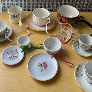 May include: A collection of miniature teacups and saucers with floral designs. The cups and saucers are white with gold trim and feature various floral patterns. Some of the cups have handles, while others are handleless. The cups and saucers are arranged on a light wood surface.