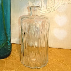May include: Two glass vases, one teal and one clear, are displayed with white and pink orchid blossoms. The teal vase is tall and cylindrical, while the clear vase is shorter with a textured design. The background is a cream-coloured fabric.