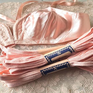 May include: A vintage lingerie set in a soft pink hue, featuring a satin bra with lace detailing and matching pink ribbon. The set is displayed on a delicate, floral-patterned lace surface, evoking a sense of classic elegance and femininity.