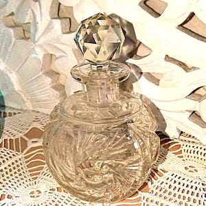 May include: A clear, vintage perfume bottle with a faceted crystal stopper. The round bottle has an etched design and sits on a white lace doily. A carved white decorative piece is in the background.