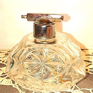 May include: A clear, faceted glass perfume bottle with a silver-coloured metal atomizer. The bottle has a geometric design and a starburst pattern on the base. The atomizer has a rectangular top.